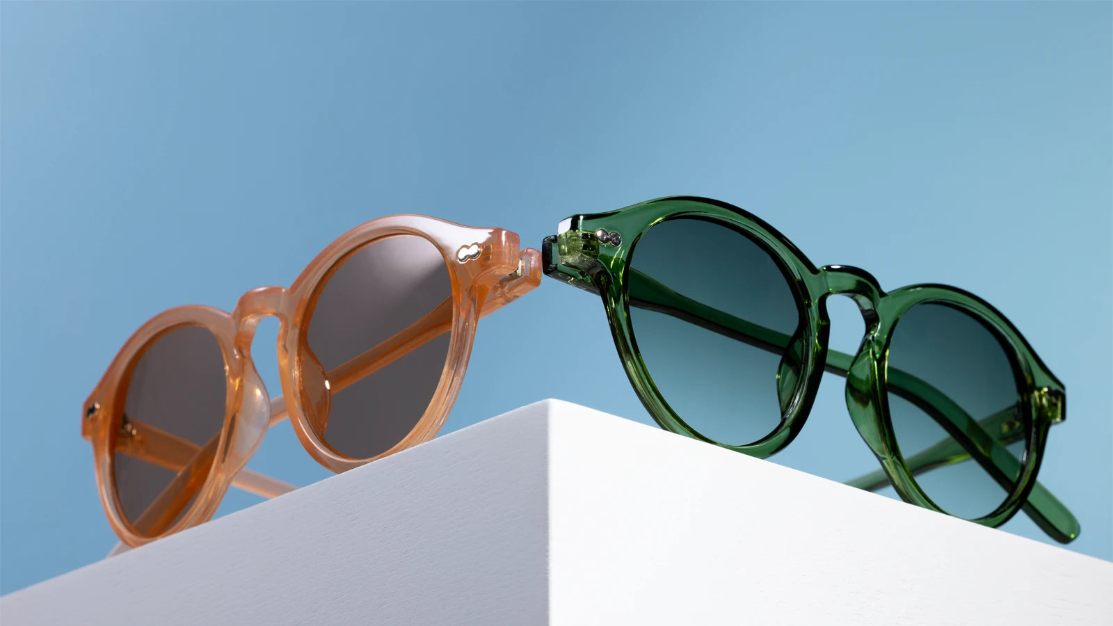 Dos pares de gafas de sol, una naranja y otra verde, sobre una superficie blanca con un fondo de cielo azul.